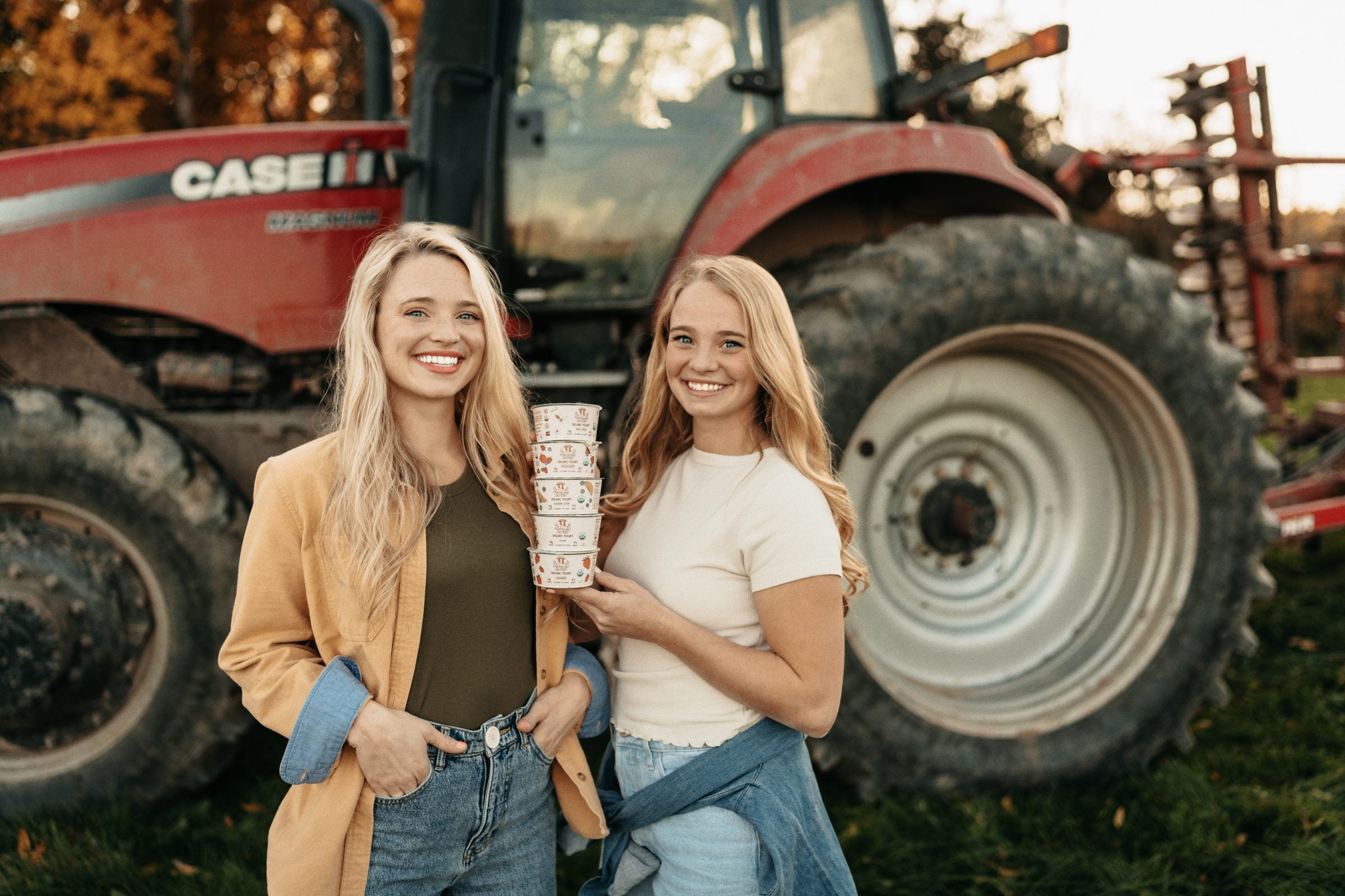 The Painterland sisters holding a stack of their organic yogurt in front of a red tractor